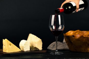 Red wine is poured into a glass, next to cheese and a serving bowl.