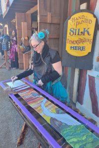 A woman wearing black and teal works on a painting next to a sign that says "Hand painted silk demonstrations here"